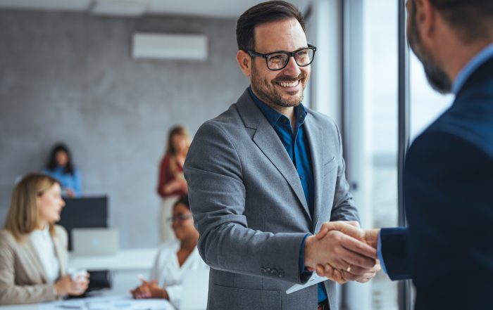 Two businessmen in suits shaking hands and smiling in a modern office, with coworkers working in the background.