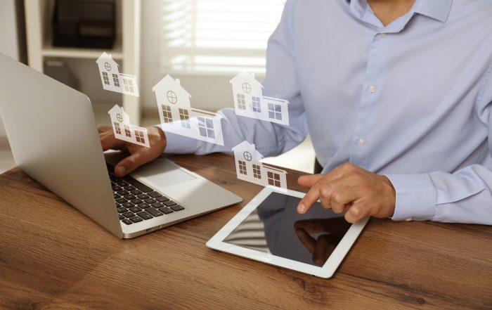 A person in a light blue shirt sits at a wooden desk using a laptop and a tablet. Floating digital house icons appear between the two devices, suggesting online real‑estate browsing or property management.