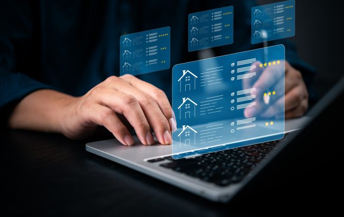 Close-up of hands using a laptop with transparent digital overlays showing house icons, property listings, feature checklists, and star ratings, representing an online real estate comparison platform.