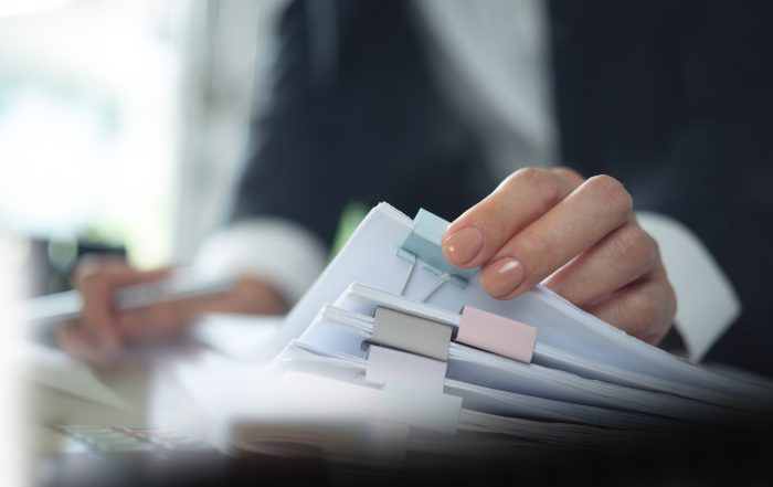 Close-up of a person reviewing a stack of paper documents with coloured binder clips and tab dividers, while writing notes at a desk in an office setting. The image highlights document organisation, records management, and administrative paperwork.
