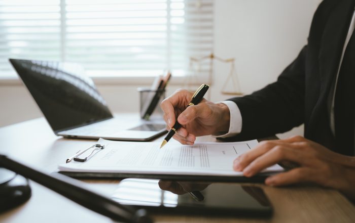 Close-up of a person in business attire signing a legal contract on a clipboard at an office desk. A laptop, tablet, and desk accessories are visible in the background, creating a professional workspace setting.