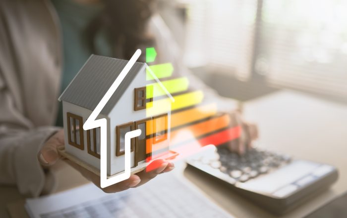 A small model house is held above a desk beside a calculator, with a colorful energy efficiency rating scale graphic overlaid on the image, illustrating home energy performance and energy-saving assessments.