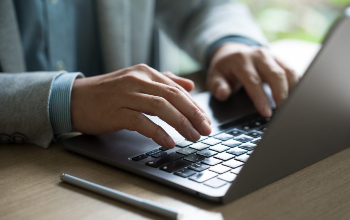 Close-up of hands typing on a laptop keyboard at a desk, with a stylus pen resting nearby. The image shows a modern workspace with natural light and a blurred background.