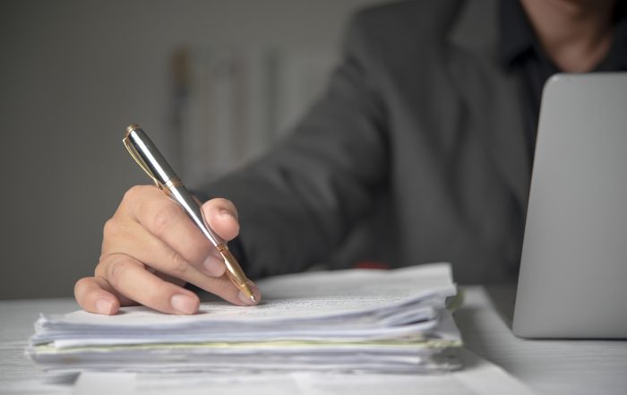Close-up of a person holding a silver and gold pen over a stack of paper documents on a desk, with an open laptop visible in the background. The image depicts reviewing, editing, or signing paperwork in an office setting.