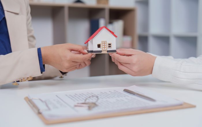 Close-up of two people exchanging a miniature house model across a desk. A clipboard with property documents and a set of keys are visible in the foreground, symbolizing a home purchase, sale, or property transfer. The background shows a softly blurred office setting.