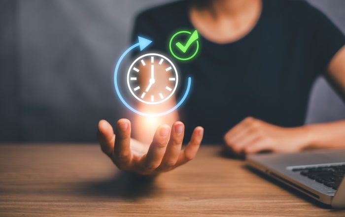 Person sitting at a desk with a laptop, holding an outstretched hand beneath a glowing holographic clock surrounded by a circular arrow and a green checkmark. The digital icons represent efficient time management, productivity, task completion, and meeting deadlines.