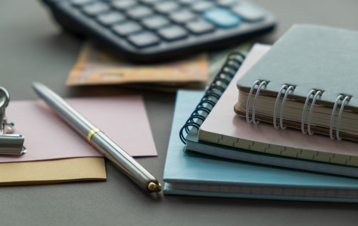 Close-up of a workspace featuring a stack of spiral-bound notebooks on a desk, with a silver pen lying across coloured notepads. A calculator and banknotes are blurred in the background, suggesting planning, budgeting or administrative work. The image is softly lit with a shallow depth of field, keeping the notebooks and pen in sharp focus.