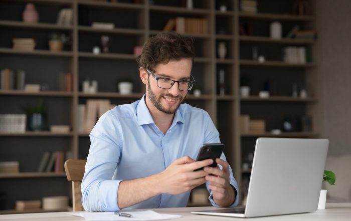 Person wearing glasses and a light blue shirt sits at a desk, holding a smartphone beside an open laptop. A large bookshelf filled with books and decorative items is visible in the background, creating a modern home-office setting.