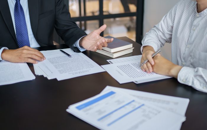 Two people seated at a meeting table reviewing printed documents. One person gestures while discussing paperwork, and the other holds a pen over a document. Reports, paperwork, and notebooks are arranged on the table in a professional office setting.