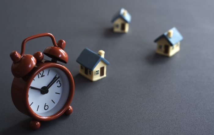 A red alarm clock and miniature houses arranged on a dark background, representing conveyancing timelines, property transactions, completion dates, and moving home schedules.